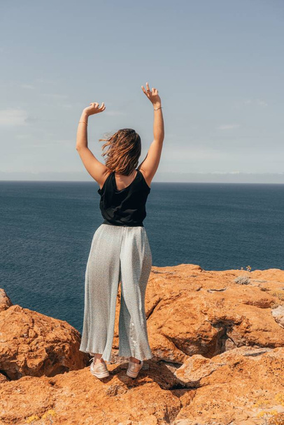A vertical shot of a European woman standing on a cliff in front of the calm blue sea with her hands in the air - Fotó, kép