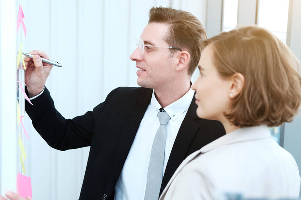 Homme d'affaires et femme d'affaires prospères écrivant de bonnes idées sur des notes collantes collées sur un mur de verre dans une réunion de bureau moderne. - Photo, image