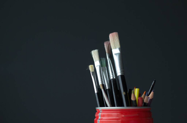 A closeup shot of paintbrushes and colored pencils in a red container on a black background - Photo, Image