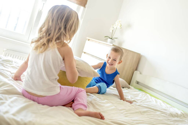Una niña y un niño sonriendo y teniendo una pelea de almohadas en una cama - Foto, Imagen