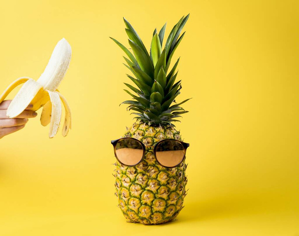 A pineapple wearing a sunglasses and a person holding a yellow banana against a yellow background - travel, summer, vacation concept - Foto, Imagen