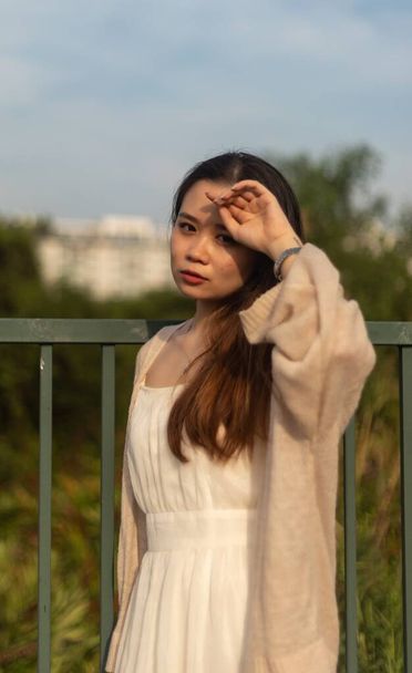 A vertical shot of an Asian female in a white dress standing with a fence on the background - Foto, imagen