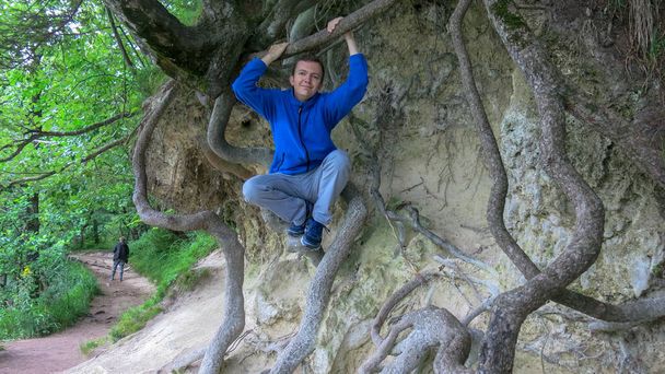 Έφηβος κάθεται στις ρίζες ενός δέντρου. Νεαρός με μπλε κουκούλα και κιλότα σκαρφαλώνει ανάμεσα στις ρίζες ενός δέντρου. Ο τύπος αναπαύεται κάτω από ένα μεγάλο δέντρο. Ο τύπος κάθεται στις ρίζες.. - Φωτογραφία, εικόνα