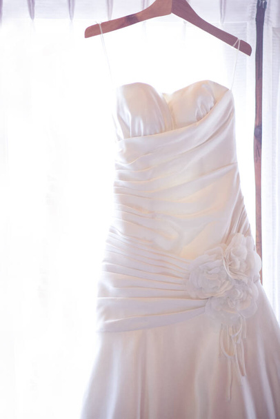 Vestido de novia colgando en el carril de la cortina cerca de ventana en la habitación
 - Foto, Imagen