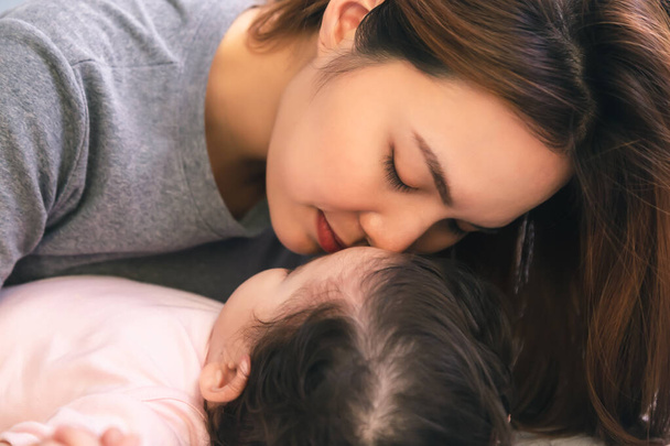 Κοντινό πλάνο νεαρή μητέρα φιλώντας το μωρό ή το κοριτσάκι της, ενώ αξιολάτρευτο κοριτσάκι κοιμάται στο κρεβάτι με αγάπη. Όμορφη ανύπαντρη μητέρα αγαπάει και φροντίζει το μωρό της τόσο πολύ. Ασιάτισσα μητέρα δώσε ένα μεγάλο φιλί - Φωτογραφία, εικόνα