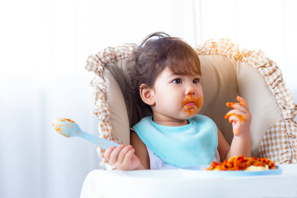 Schattige hongerige peuter meisje of baby eten heerlijke spaghetti voedsel met tomatensaus op kinderstoel. Grappig schattig baby meisje krijgen honger en eten alleen. Dochter wordt vies, kijk weg. - Foto, afbeelding