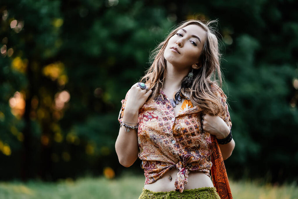 Beautiful young woman wearing bohostyle clothes posing in the rays of the evening sun, sunset. Boho style fashion, female wearing silver jewelry having fun in park outdoors. - Photo, Image