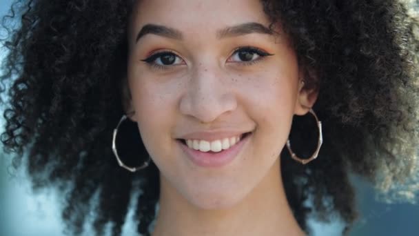Schöne selbstbewusste junge Afrikanerin lacht. Glücklicher ethnischer Teenager mit braunen Augen, lockigen Afro-Frisuren, runden Ohrringen, leichtem Make-up und Sommersprossen lächelt und zeigt ihre gesunden weißen Zähne. - Filmmaterial, Video
