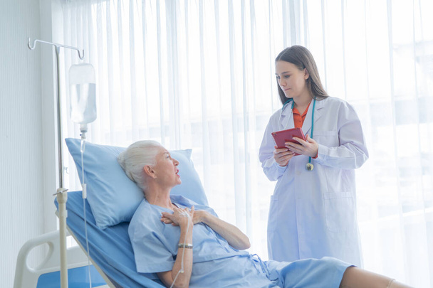 Happy woman doctor talking and using tablet to sick old female senior elderly patient lying in bed in hospital ward room in medical, technology and healthcare treatment concept. Caucasian people. - Photo, Image