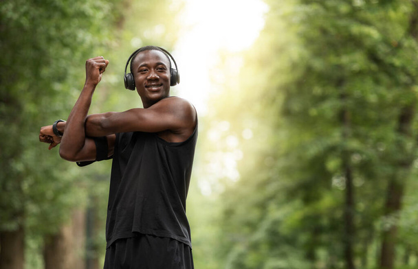 Веселый африканский спортсмен, тренирующийся в парке, вытягивающий руки
 - Фото, изображение