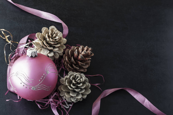 Weihnachtlicher Hintergrund. Drei Tannenzapfen und eine rosa gläserne Weihnachtskugel auf schwarzem Hintergrund. Festliche Stimmung. Kopierraum. - Foto, Bild