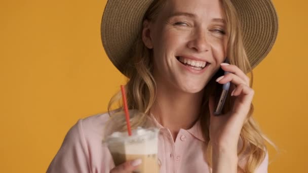 Chica rubia bastante sonriente en sombrero bebiendo café helado felizmente hablando en el teléfono inteligente sobre fondo colorido
 - Metraje, vídeo