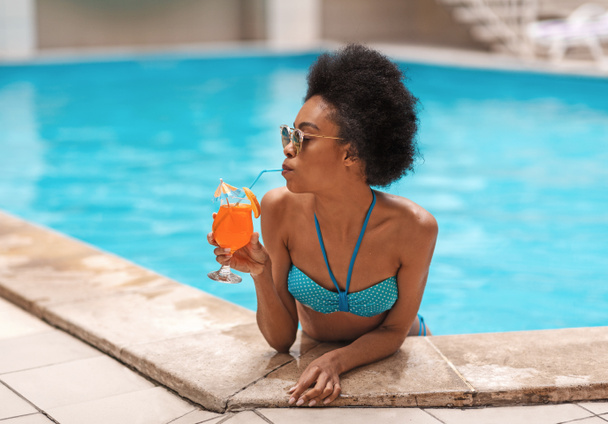 sexy czarny dziewczyna w niebieski bikini sącząc smaczny letni koktajl w odkryty basen - Zdjęcie, obraz