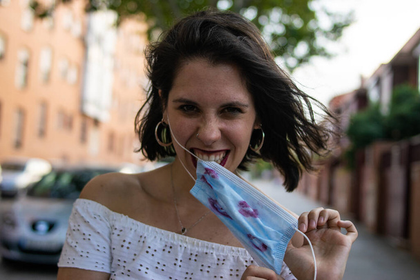 Foto de una mujer joven y atractiva con una máscara facial azul desechable cubierta con lápiz labial con patrón de labios y aspecto feliz - Foto, Imagen
