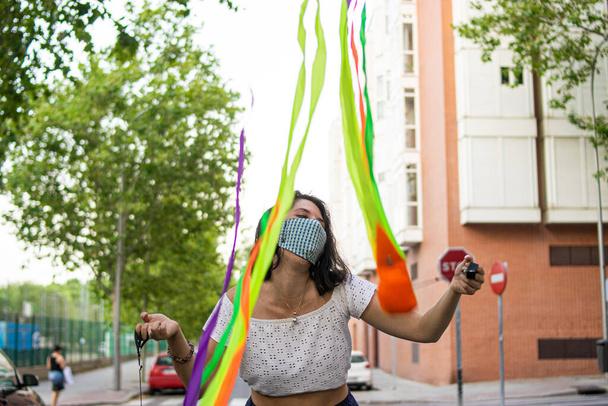 Фото молодой и привлекательной женщины, одетой в маску многоразового использования и играющей в жонглирование с красочными полосками - Фото, изображение