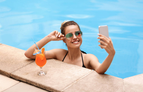 Lovely Caucasian girl taking selfie on smartphone in pool at tropical resort - Фото, изображение