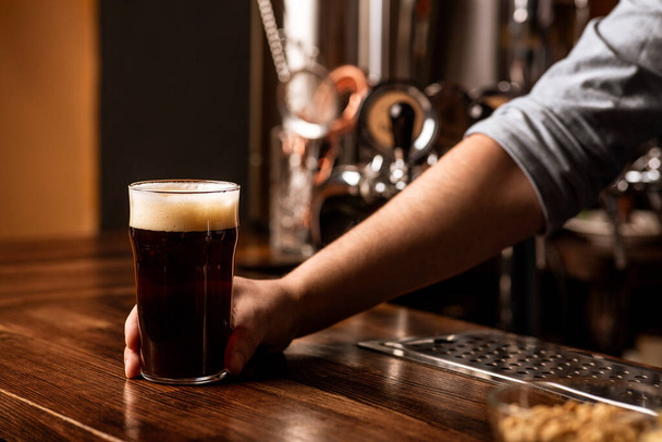 Tuoretta tummaa olutta, jossa on vaahtoa pubissa. Baarimikko antaa lasi tumma juoma vaahto baaritiskillä - Valokuva, kuva