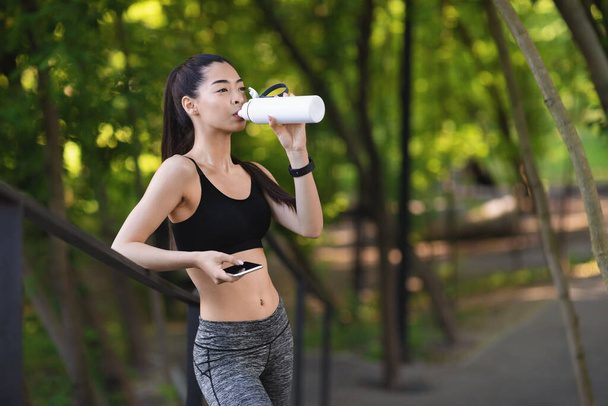 Happy young asian girl drinking water after jogging in park, exercising outdoors - Photo, Image