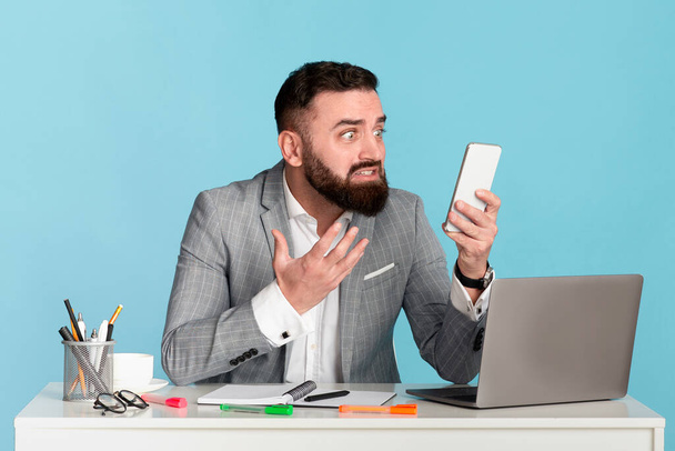 Pissed office employee staring at his mobile phone at workplace over blue background - Фото, изображение