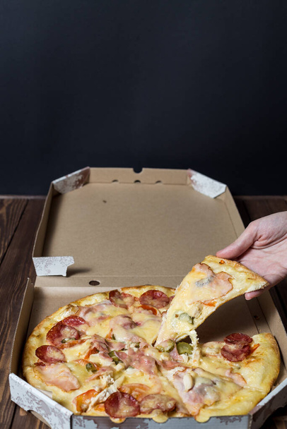 Женская рука берет кусочек пиццы. пицца в открытой картонной коробке на деревянном столе. фаст-фуд рекламный плакат. - Фото, изображение