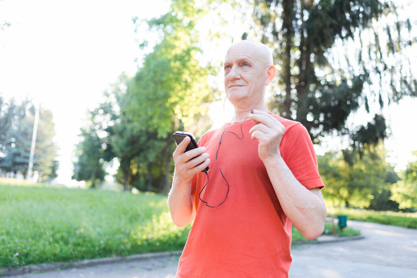 Hombre viejo sonriente en ropa casual y auriculares blancos escuchando música desde su teléfono inteligente en el parque de la ciudad en un día soleado
. - Foto, Imagen