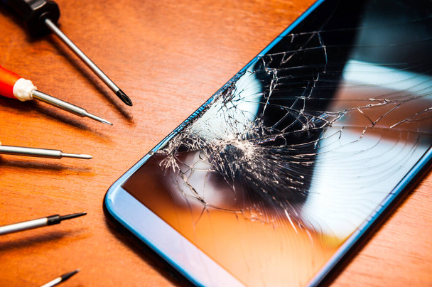 A broken mobile phone with a broken screen with tools, screwdrivers on a wooden table. Electronics repair shops. - Zdjęcie, obraz