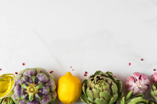 Carciofi freschi maturi, limone e aglio con olio d'oliva e pepe sul tavolo di marmo bianco. Sano concetto di cibo vegano. Piatto. vista dall'alto con spazio di copia - Foto, immagini