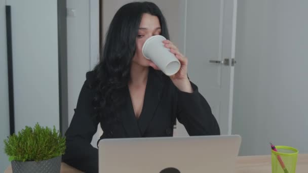 Vrouw gebruikt laptop terwijl ze aan haar bureau zit en koffie drinkt. Jonge Europese zakenvrouw zit in het kantoor en werkt aan computersoftware - Video