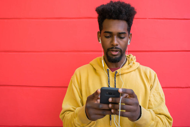Portrait of young afro man using his mobile phone with earphones against red wall. Technology and lifestyle concept. - Photo, Image