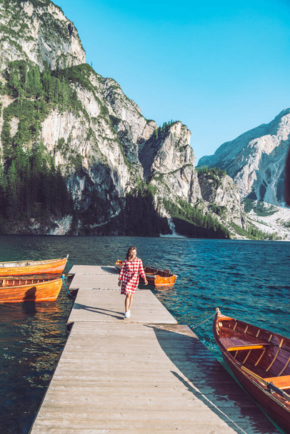 woman walking by mountain lake pier with wooden boats in red dress - 写真・画像