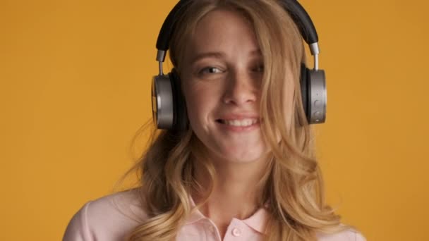 Portrait of pretty charming blond girl dreamily listening music in headphones over colorful background - Felvétel, videó