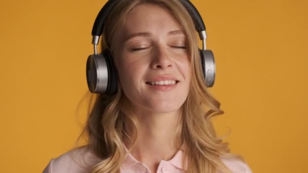 Portrait of beautiful smiling blond girl dreamily listening music in headphones over colorful background - Felvétel, videó