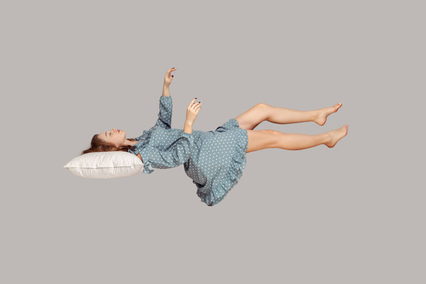 Sleeping beauty floating in air. Relaxed girl in vintage ruffle dress keeping eye closed, lying on pillow levitating, flying in dream with hands up to catch. studio shot isolated on gray background - 写真・画像