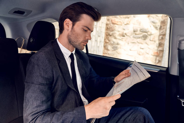 Reading latest news. Handsome young man in full suit reading a newspaper while sitting in the car. - Zdjęcie, obraz
