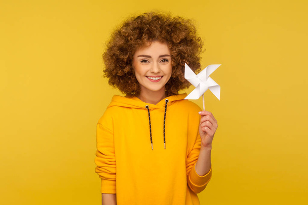 Origami hand mill. Portrait of happy curly-haired woman in urban style hoodie smiling carefree and holding paper windmill, pinwheel toy on stick. indoor studio shot isolated on yellow background - Foto, afbeelding