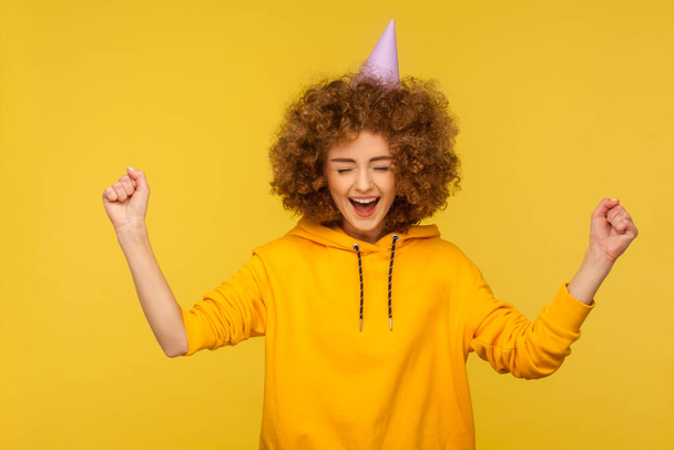 Portrait of overjoyed curly-haired hipster woman with funny cone hat raising hands, screaming with happiness and hanging out at party, celebrating holiday. studio shot isolated on yellow background - Foto, Bild