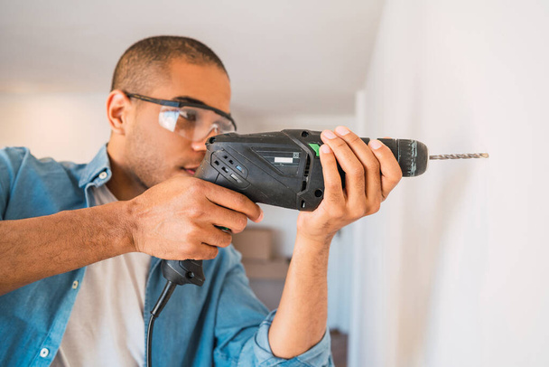 Portrait of young latin man with a electric drill and making hole in wall. Interior design and home renovation concept. - 写真・画像