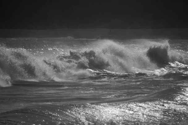 A stormy sea with gale force winds and the high tide creates huge waves which crash on the rocky beach. The setting sun glints across the water and the spray creates a mist across the water.  - Photo, Image