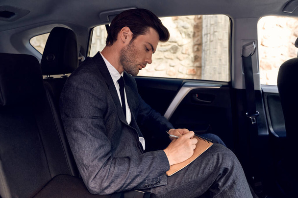 Always In Touch Concept. Young manager writing in notebook sitting in back seat of car. - Foto, Bild