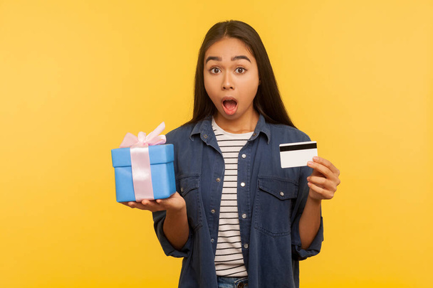 Wow gift shopping! Portrait of amazed girl in denim shirt holding present box, credit card and looking surprised, shocked by purchase, cashback and bank loan. studio shot isolated on yellow background - Photo, image