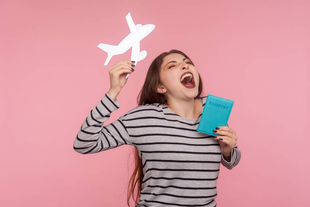 Ура, я отправляюсь в путешествие! Портрет эйфории взволнованной восторженной женщины, кричащей от счастья, держащей паспорт и бумажный самолет, радующейся путешествиям. студия съемки изолированы на розовом фоне
 - Фото, изображение
