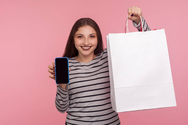 Online store app. Portrait of happy woman in striped sweatshirt holding shopping bag and cell phone, showing mobile device with blank mock up for advertise. indoor studio shot, yellow background - Photo, Image