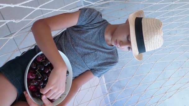 Лето, каникулы, концепция здорового питания. Мальчик ест сладкие свежие вишни с тарелки, раскачиваясь в гамаке. Видеозапись с вертикальной ориентацией экрана. 4k - Кадры, видео