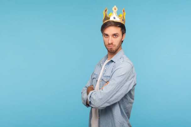I'm king! Portrait of confident ambitious man wearing golden crown, looking with arrogance, declaring his authority and leadership, superior privileged status. studio shot isolated on blue background - Foto, Bild