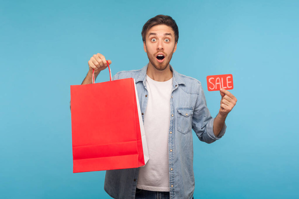 Portrait of surprised shopper man in denim shirt holding Sale inscription and packages with blank space for advertise text, discount in store, expressing shock. studio shot isolated on blue background - Foto, imagen