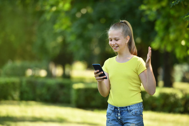 Smiling girl child received a joyful message in a smartphone on a background of green trees in a park - Fotoğraf, Görsel