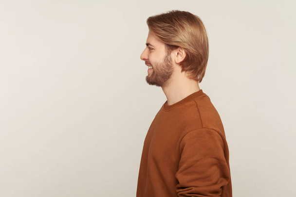 Profile of happy cheerful handsome man with neat hair and beard wearing sweatshirt standing, smiling to side copy space, feeling optimistic positive. indoor studio shot isolated on gray background - Photo, Image