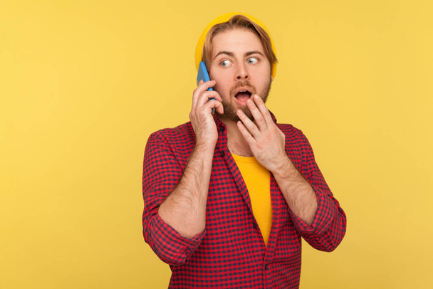 Surprised trendy guy in hipster beanie hat and checkered shirt calling, expressing shock emotions while talking mobile phone, hearing unbelievable news on telephone. indoor studio shot isolated - Photo, Image