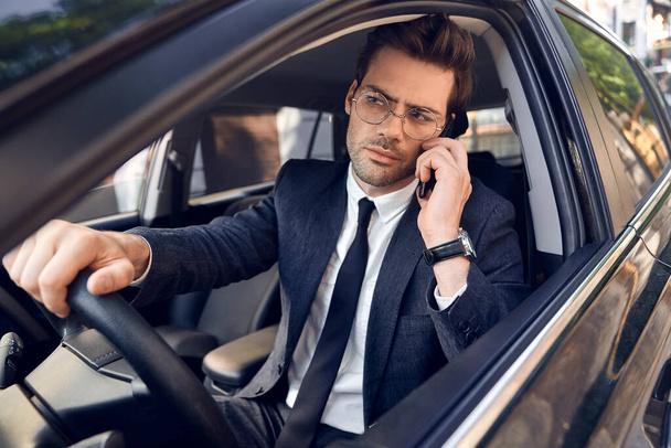 Nice business talk. Handsome young businessman talking on his smart phone and smiling while sitting on the front seat. - Fotoğraf, Görsel