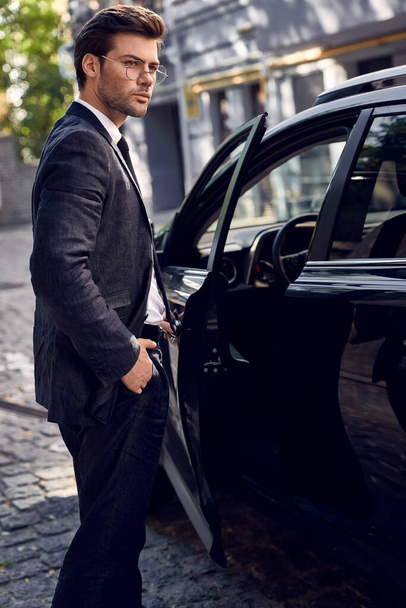 Πολυτελές στυλ. Όμορφος νεαρός επιχειρηματίας μπαίνει στο αυτοκίνητό του, ενώ στέκεται έξω. - Φωτογραφία, εικόνα